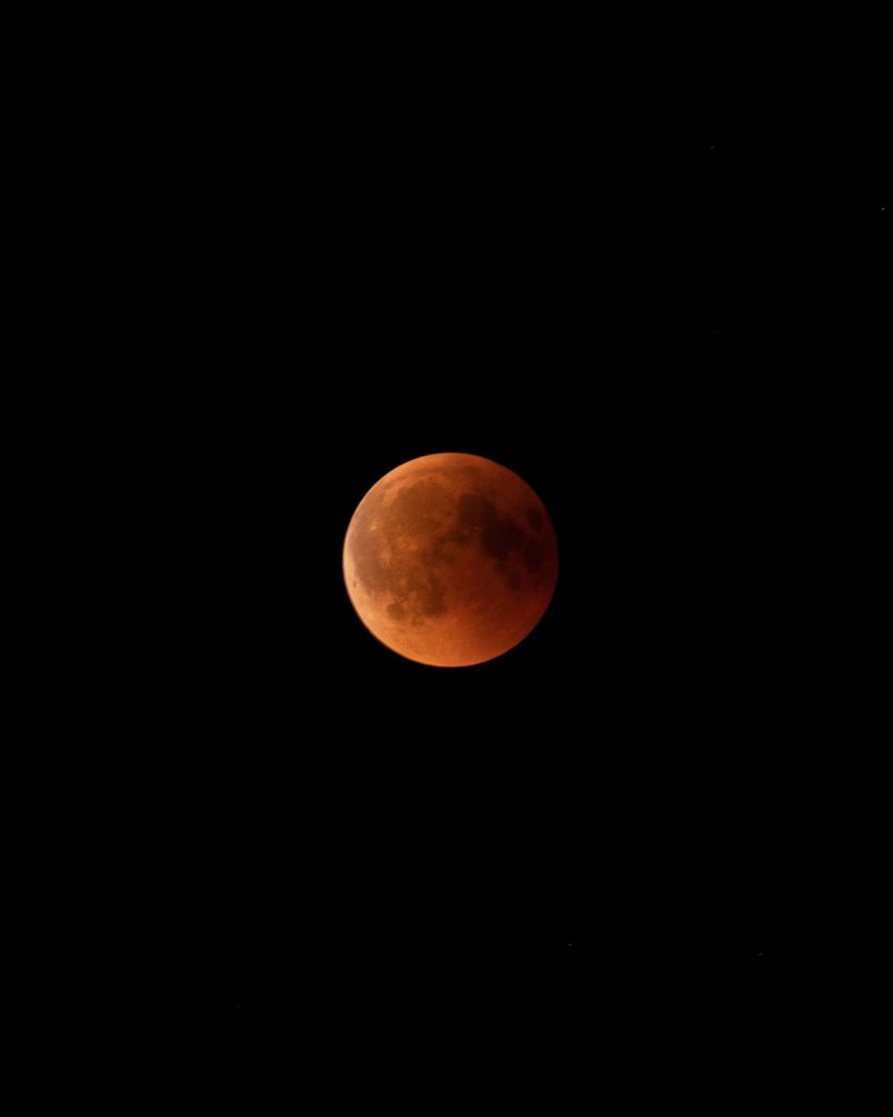 L’image montre une éclipse lunaire partielle dans un ciel noir. La Lune apparaît rougeâtre avec des zones d’ombre visibles. Aucun autre élément n’est présent, mettant en valeur l’astre.