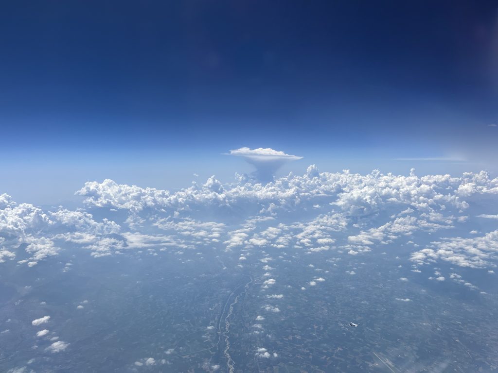 Une superbe vue aérienne avec un ciel bleu profond parsemé de nuages cotonneux. Un nuage en forme d'enclume se distingue à l'horizon, flottant majestueusement au-dessus d'un paysage terrestre à peine visible sous une fine brume.