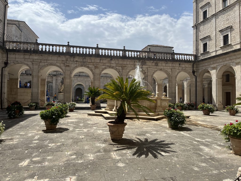 Une élégante cour intérieure baignée de soleil, ornée d'une fontaine centrale et entourée d'arches en pierre. Des plantes en pots et des fleurs colorées ajoutent une touche de vie à cette architecture historique et apaisante.