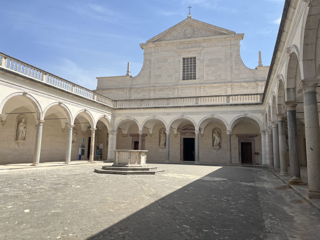 Une cour paisible d'un monastère, bordée d'arcades et de colonnes en pierre. En son centre, un puits en marbre et des statues sculptées ornent les murs. L'atmosphère sereine est renforcée par la lumière douce du jour et l'ombre des colonnades.