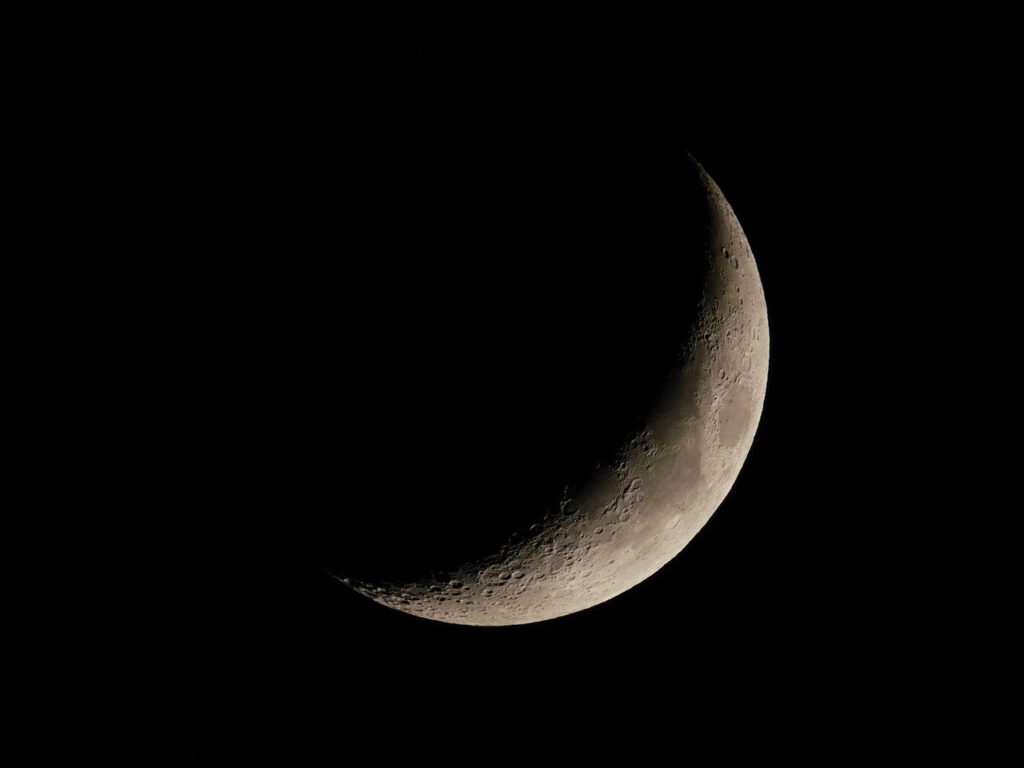 Photo d'un croissant de lune dans un ciel nocturne profond et sombre. La surface lunaire révèle des détails texturés avec des cratères visibles, illuminée par la lumière solaire sur une partie de l'astre, tandis que le reste demeure plongé dans l'ombre.