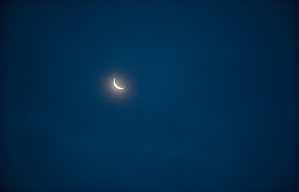 Dans cette image, un fin croissant de lune illumine doucement le ciel nocturne, entouré d'une lueur diffuse. L'immensité du bleu profond qui l'entoure renforce la sensation de calme et de mystère. La simplicité de cette scène céleste invite à la contemplation et à la rêverie.