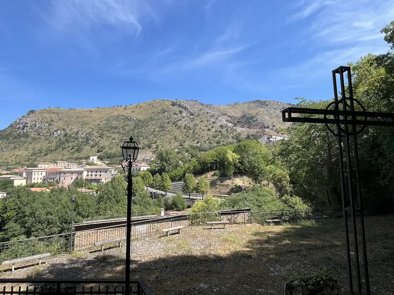 Village pittoresque niché au pied de montagnes et collines verdoyantes, avec une croix métallique et un lampadaire au premier plan.